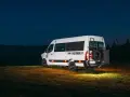 Biały kamper Renault Master z kołem zapasowym z tyłu, zaparkowany na łące nocą.