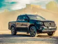 Czarny Mercedes pickup z przyciemnianymi szybami, zaparkowany na piaszczystym podłożu, w tle przemysłowy budynek i unoszący się pył.