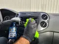 Ręce w czarnych rękawiczkach wykonują polerowanie plastiku na desce rozdzielczej samochodu, używając zielonej ściereczki i płynu.