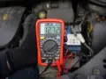Ręka w czarnej rękawiczce trzyma multimetr pokazujący 12.72V. To idealne minimalne napięcie akumulatora do uruchomienia.