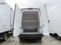 Biała izoterma z otwartymi drzwiami tylnymi, widoczne półki i wnętrze. Obok inne białe zabudowy.