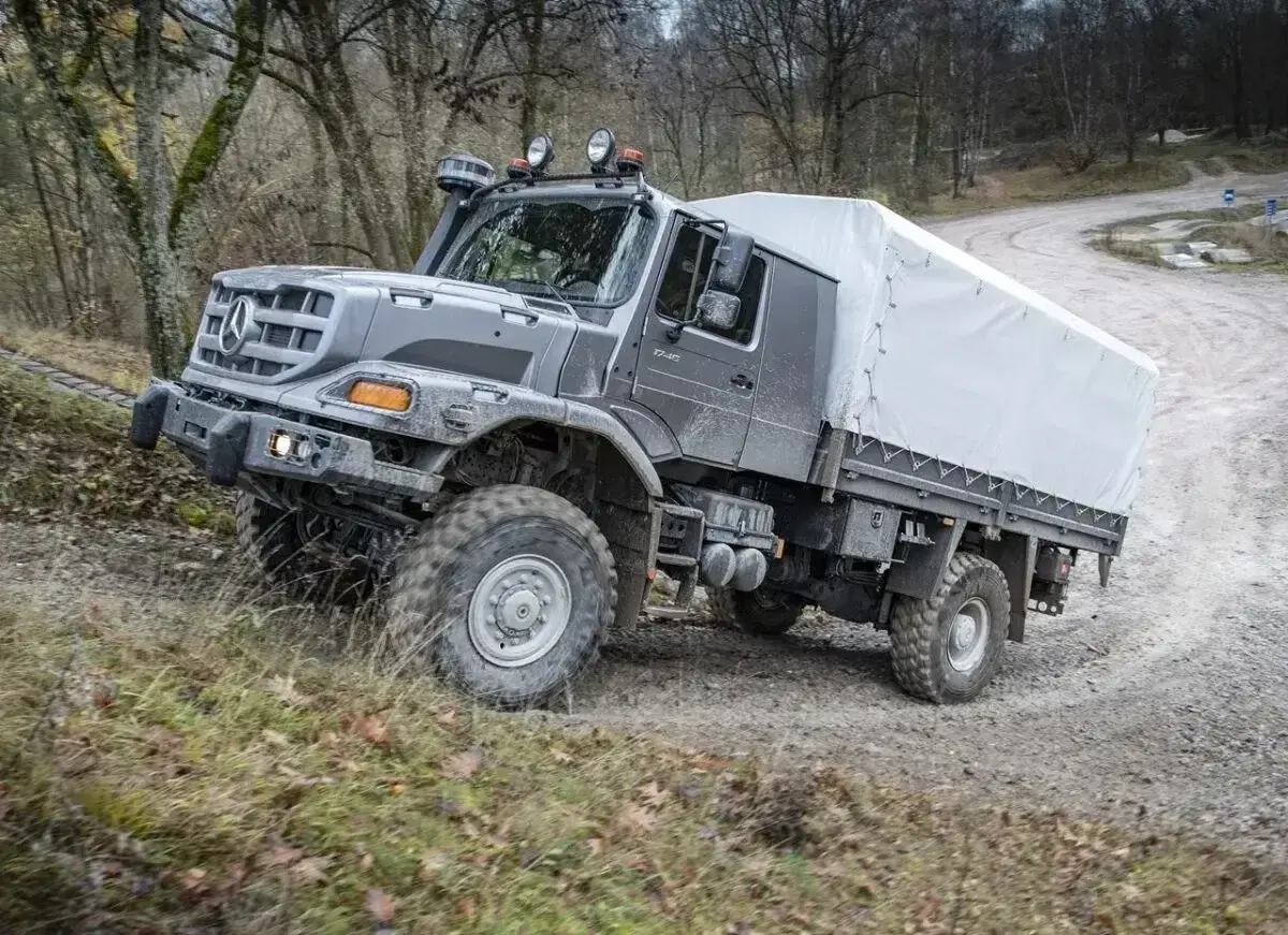 Szary Mercedes Zetros z plandeką pokonuje trudny teren leśnej drogi.