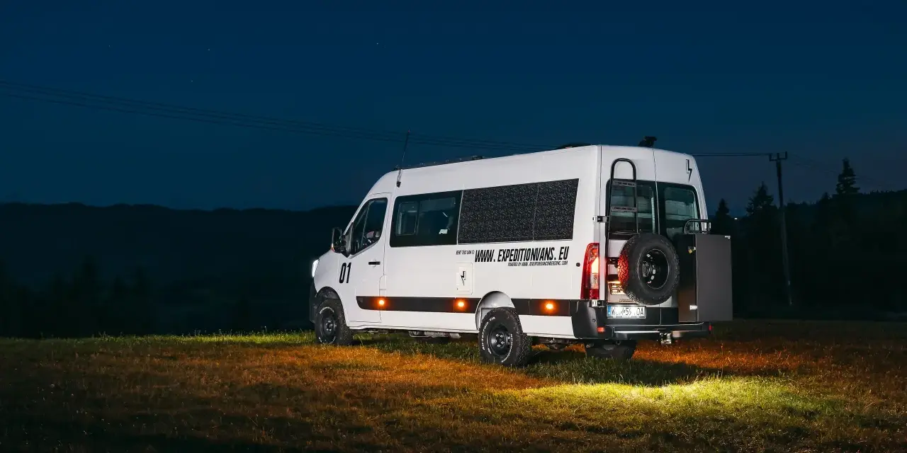 Biały kamper Renault Master z kołem zapasowym z tyłu, zaparkowany na łące nocą.