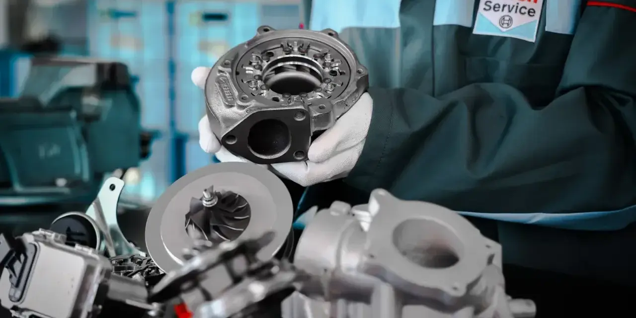 Mechanik w białych rękawiczkach prezentuje części turbosprężarki, sugerując domowe ustawienie sztangi turbiny.