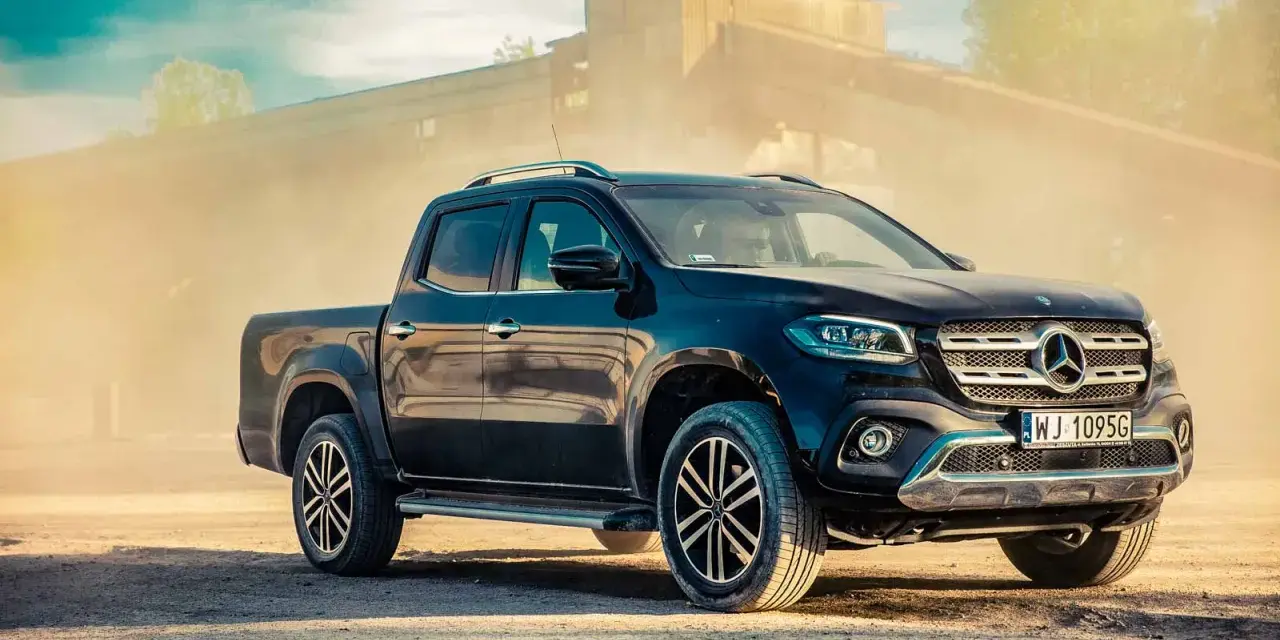 Czarny Mercedes pickup z przyciemnianymi szybami, zaparkowany na piaszczystym podłożu, w tle przemysłowy budynek i unoszący się pył.