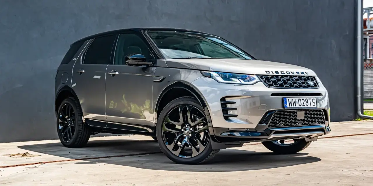Srebrny Land Rover Discovery Sport z czarnym dachem, zaparkowany przed szarą ścianą.