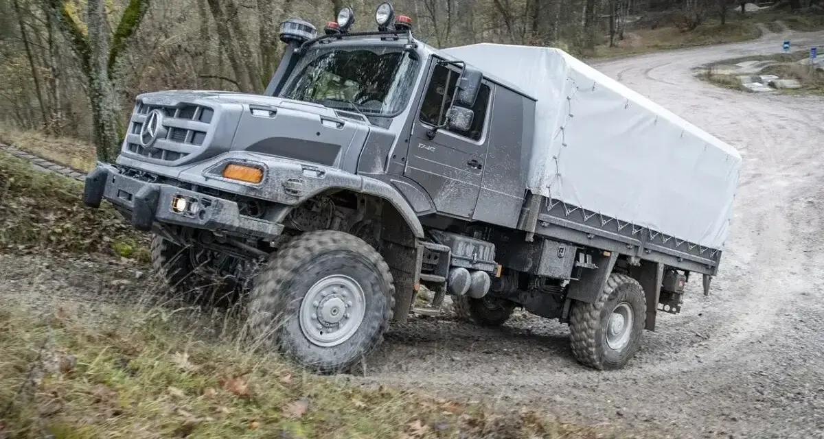 Szary Mercedes Zetros z plandeką pokonuje trudny teren leśnej drogi.