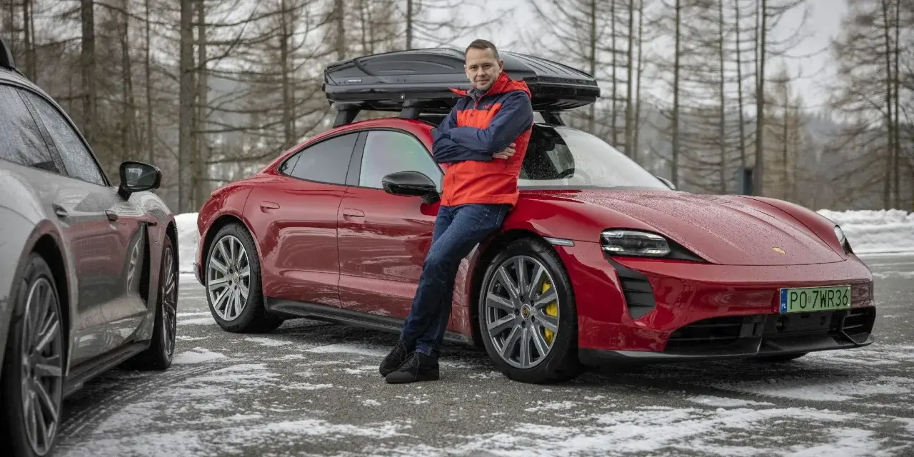 Mężczyzna w czerwonej kurtce opiera się o czerwonego Porsche Taycan z boxem dachowym.