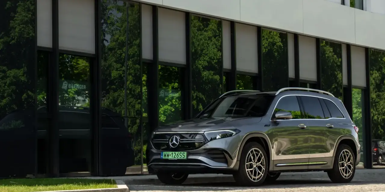 Nowy Mercedes 4x4, elegancki SUV, zaparkowany przed nowoczesnym budynkiem z zielonymi odbiciami drzew w szybach.