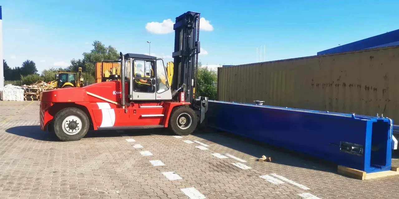 Czerwony wózek widłowy przygotowuje się do przeładunku długiego, niebieskiego elementu. To przykład sprawnego cross dockingu.