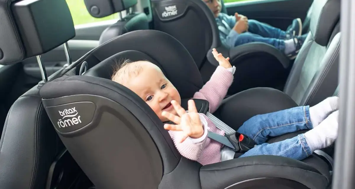 Uśmiechnięte niemowlę w foteliku Britax Römer, obok starsze dziecko. Od kiedy dziecko może jeździć z przodu? To pytanie nurtuje wielu rodziców.