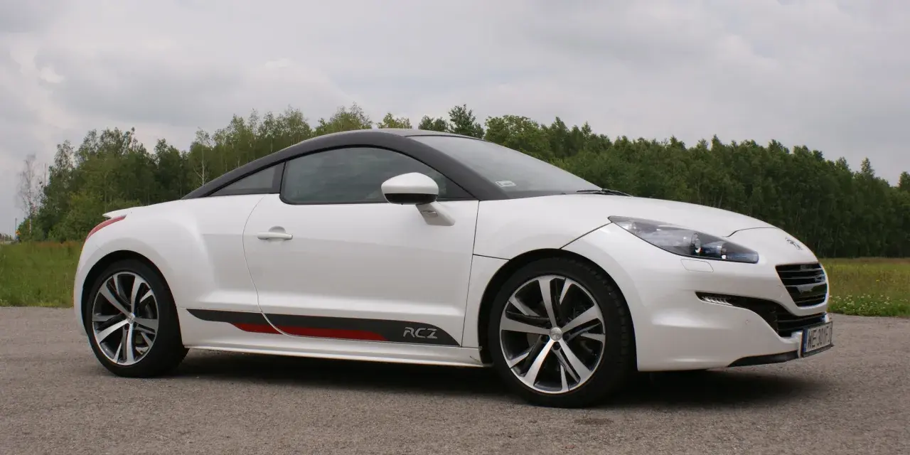Biały Peugeot RCZ z czarnym dachem i sportowymi akcentami na progu, zaparkowany na tle zielonego lasu.