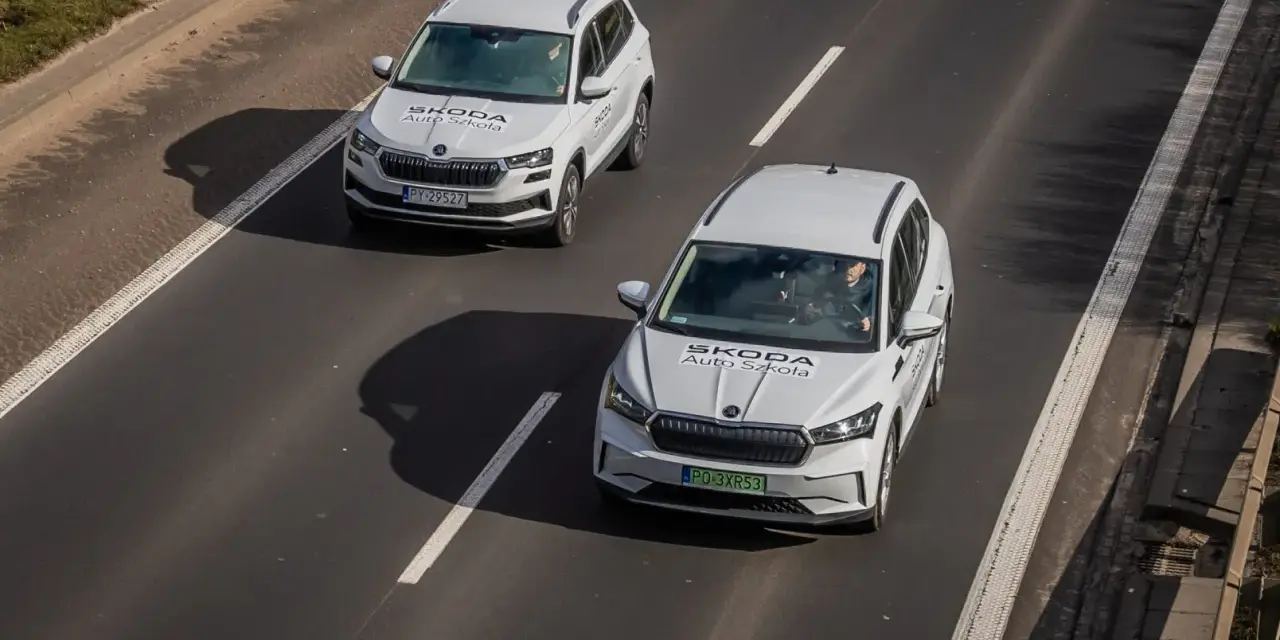 Dwa białe samochody Skoda Auto Szkoła jadą po drodze, gdzie obowiązuje ruch lewostronny.
