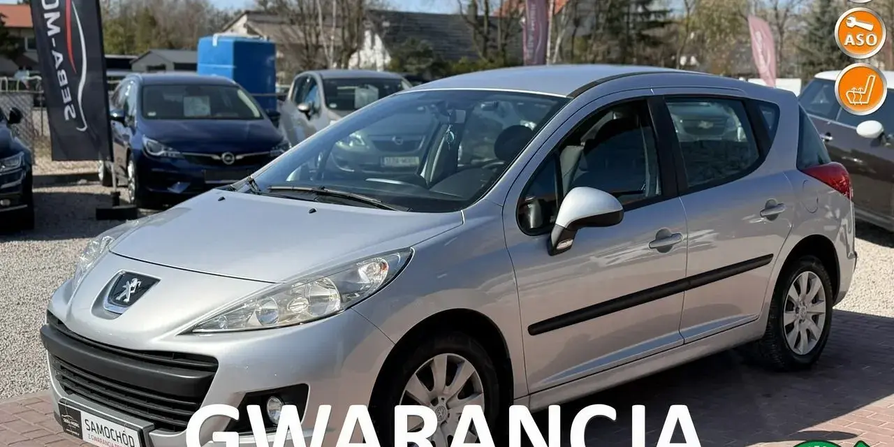 Srebrny Peugeot 207 1.4 benzyna na placu samochodowym, z napisem "GWARANCJA".