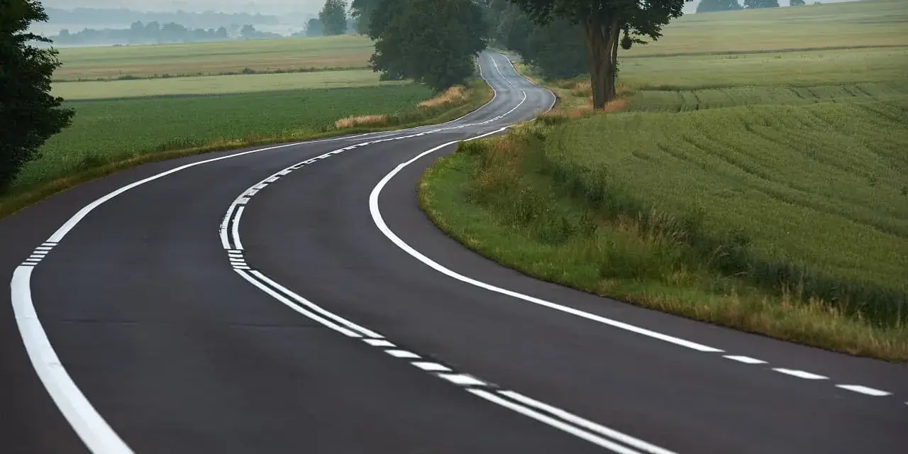 Zakręt drogi z podwójną ciągłą linią, otoczony polami i drzewami. Kierowca rozważa wyprzedzanie na podwójnej ciągłej.