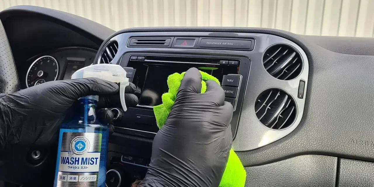 Ręce w czarnych rękawiczkach wykonują polerowanie plastiku na desce rozdzielczej samochodu, używając zielonej ściereczki i płynu.