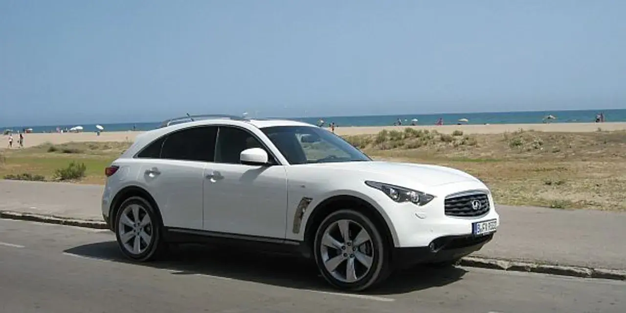 Białe infiniti auto zaparkowane przy plaży, z widokiem na morze i ludzi odpoczywających na piasku.
