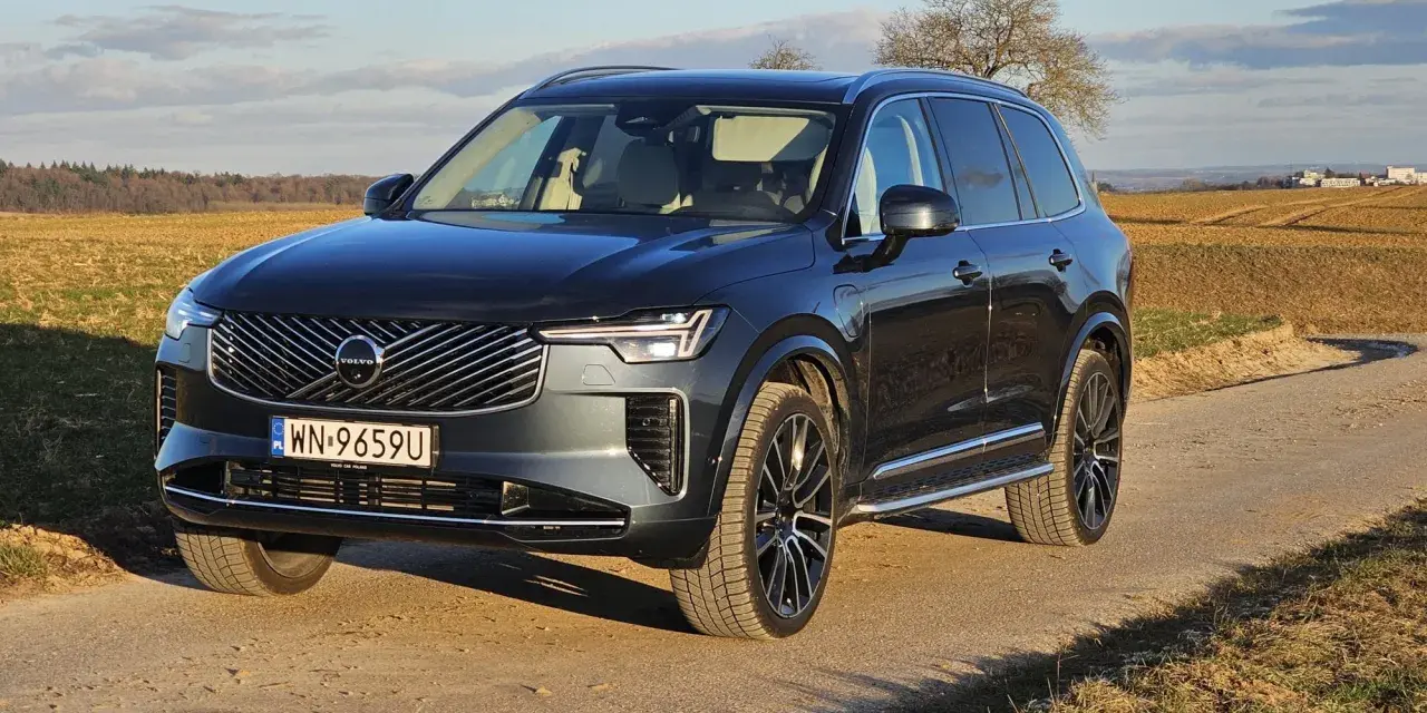 Ciemnoszary Volvo XC90 na polnej drodze, z widokiem na pola i drzewa w tle.
