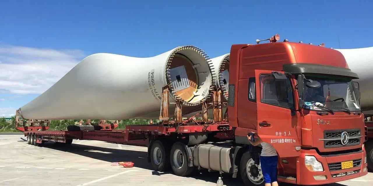 Pomarańczowy tir przewozi gigantyczne łopaty turbin wiatrowych. Kierowca przygotowuje się do dalszej podróży.