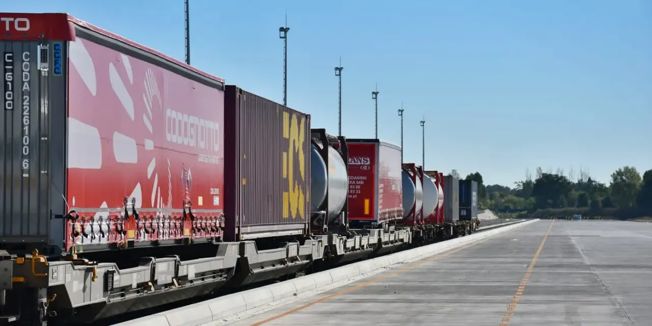 Długi pociąg towarowy z kontenerami, w tym czerwonymi z logo COSCO i żółtymi, czeka na dalszy transport kolejowy.