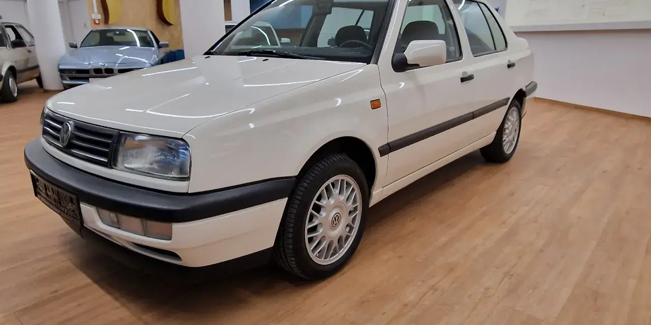 Biały Volkswagen Vento z lat 90. stoi na drewnianej podłodze w salonie samochodowym.