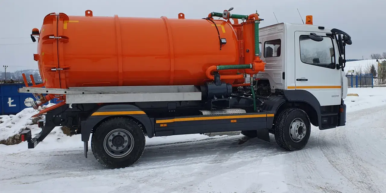 Pomarańczowy pojazd asenizacyjny z beczką i zielonymi wężami stoi na zaśnieżonej drodze.