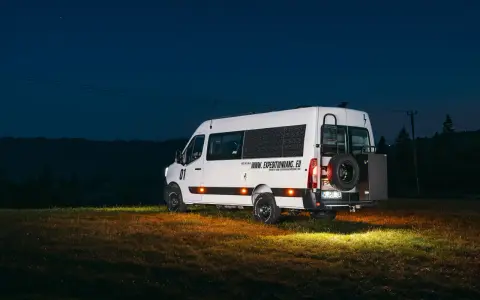 Biały kamper Renault Master z kołem zapasowym z tyłu, zaparkowany na łące nocą.