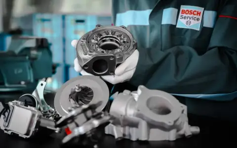 Mechanik w białych rękawiczkach prezentuje części turbosprężarki, sugerując domowe ustawienie sztangi turbiny.