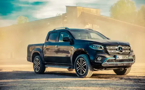 Czarny Mercedes pickup z przyciemnianymi szybami, zaparkowany na piaszczystym podłożu, w tle przemysłowy budynek i unoszący się pył.