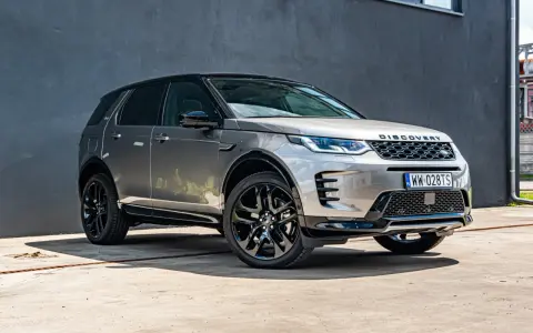 Srebrny Land Rover Discovery Sport z czarnym dachem, zaparkowany przed szarą ścianą.