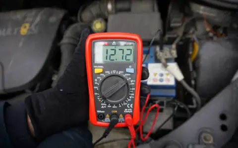 Ręka w czarnej rękawiczce trzyma czerwony multimetr z wyświetlaczem 12.72V, na tle silnika samochodu.