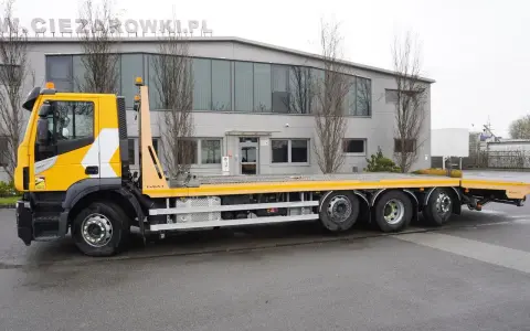Żółty Iveco Stralis z platformą transportową stoi przed budynkiem.