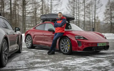 Mężczyzna w czerwonej kurtce opiera się o czerwonego Porsche Taycan z boxem dachowym.
