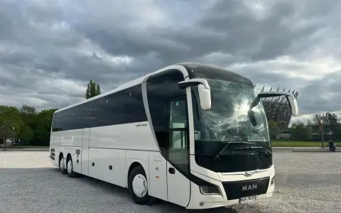 Biały MAN Lion's Coach, nowoczesny autokar turystyczny, zaparkowany na żwirowym placu pod pochmurnym niebem.