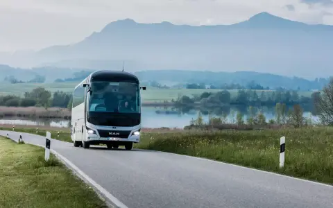 Biały **bus** marki **MAN** jedzie krętą drogą nad jeziorem, w tle góry.
