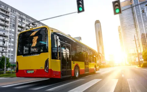 Żółto-czerwony autobus elektryczny Yutong jedzie przez miasto w blasku zachodzącego słońca.
