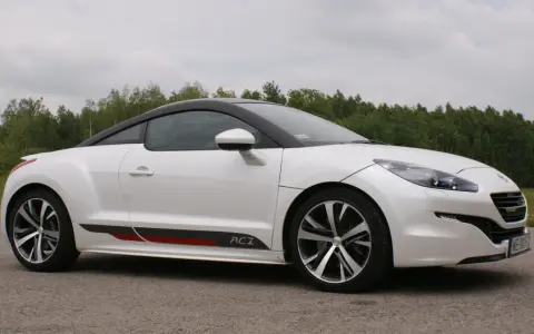 Biały Peugeot RCZ z czarnym dachem i sportowymi akcentami na progu, zaparkowany na tle zielonego lasu.