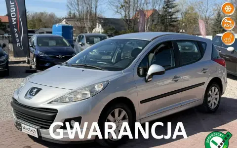 Srebrny Peugeot 207 1.4 benzyna na placu samochodowym, z napisem "GWARANCJA".