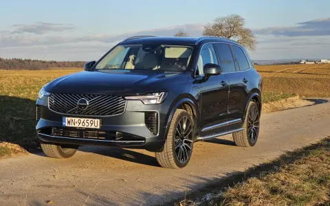 Ciemnoszary Volvo XC90 na polnej drodze, z widokiem na pola i drzewa w tle.