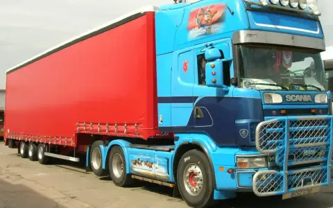 Naczepa jumbo w jaskrawym kolorze czerwonym, ciągnięta przez niebieską Scanię z malowidłami.