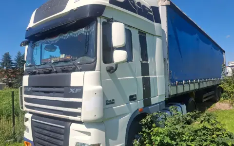 Ciężarówka DAF XF 105 z niebieską plandeką stoi na trawie, częściowo zasłonięta przez krzaki.