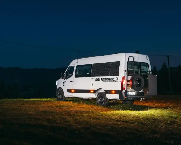 Biały kamper Renault Master z kołem zapasowym z tyłu, zaparkowany na łące nocą.