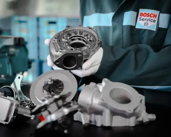 Mechanik w białych rękawiczkach prezentuje części turbosprężarki, sugerując domowe ustawienie sztangi turbiny.