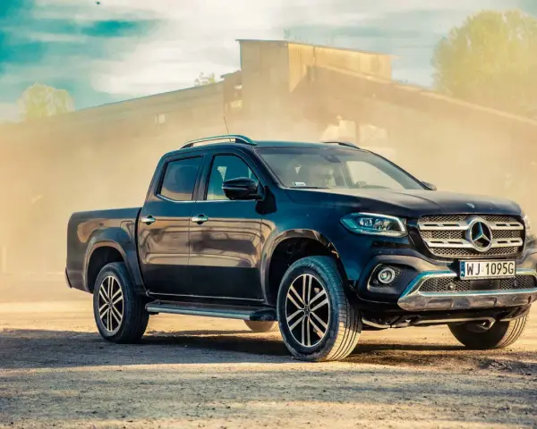 Czarny Mercedes pickup z przyciemnianymi szybami, zaparkowany na piaszczystym podłożu, w tle przemysłowy budynek i unoszący się pył.
