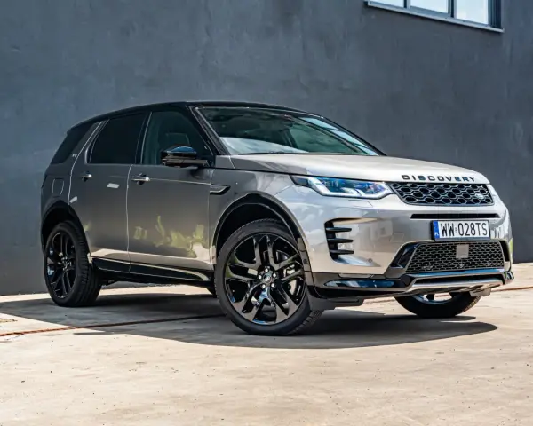 Srebrny Land Rover Discovery Sport z czarnym dachem, zaparkowany przed szarą ścianą.