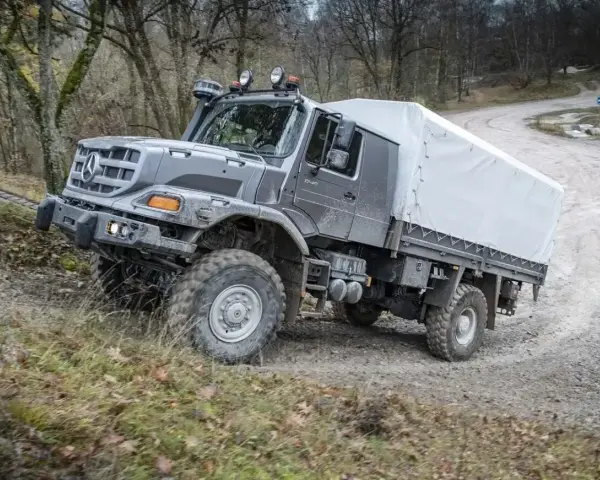 Szary Mercedes Zetros z plandeką pokonuje trudny teren leśnej drogi.