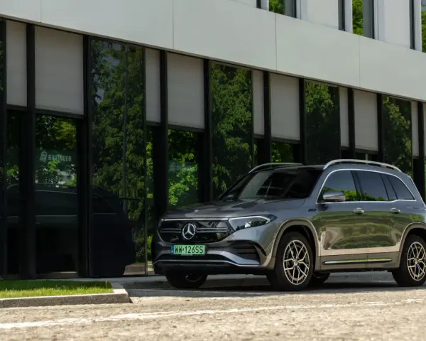 Nowy Mercedes 4x4, elegancki SUV, zaparkowany przed nowoczesnym budynkiem z zielonymi odbiciami drzew w szybach.