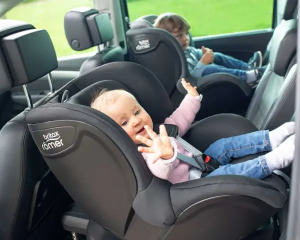 Uśmiechnięte niemowlę w foteliku Britax Römer, obok starsze dziecko. Od kiedy dziecko może jeździć z przodu? To pytanie nurtuje wielu rodziców.