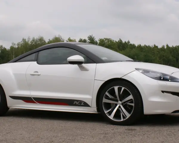 Biały Peugeot RCZ z czarnym dachem i sportowymi akcentami na progu, zaparkowany na tle zielonego lasu.
