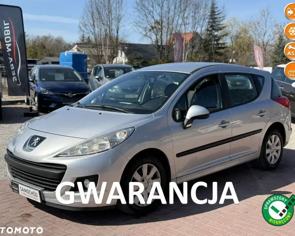 Srebrny Peugeot 207 1.4 benzyna na placu samochodowym, z napisem "GWARANCJA".